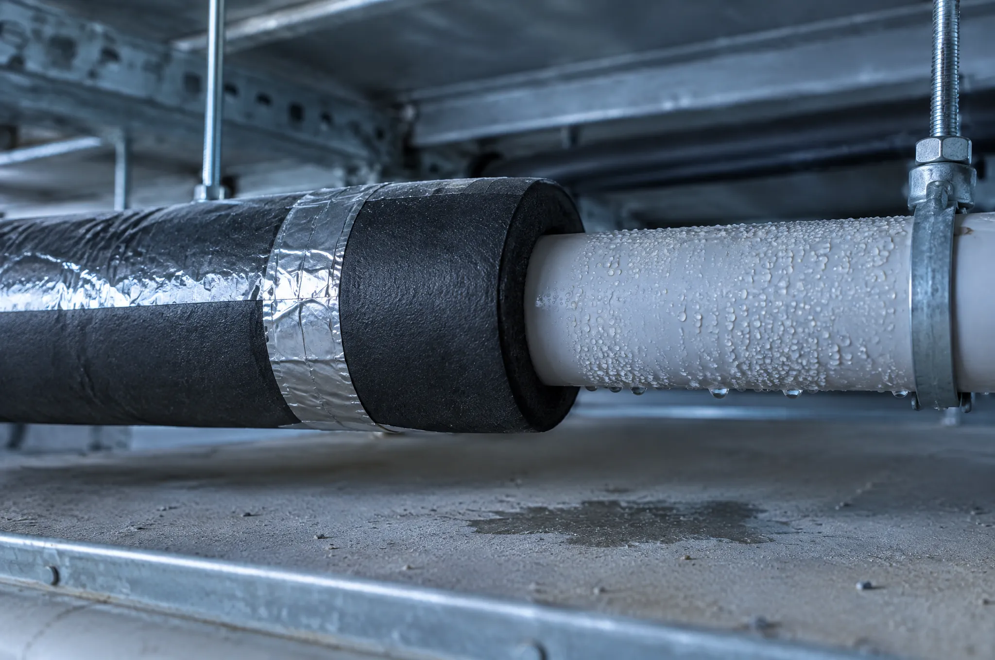 A chilled water pipe where the closed-cell insulation and its taped foil vapour barrier stop: the bare section beyond is beaded with condensation and a damp mark has formed on the surface below