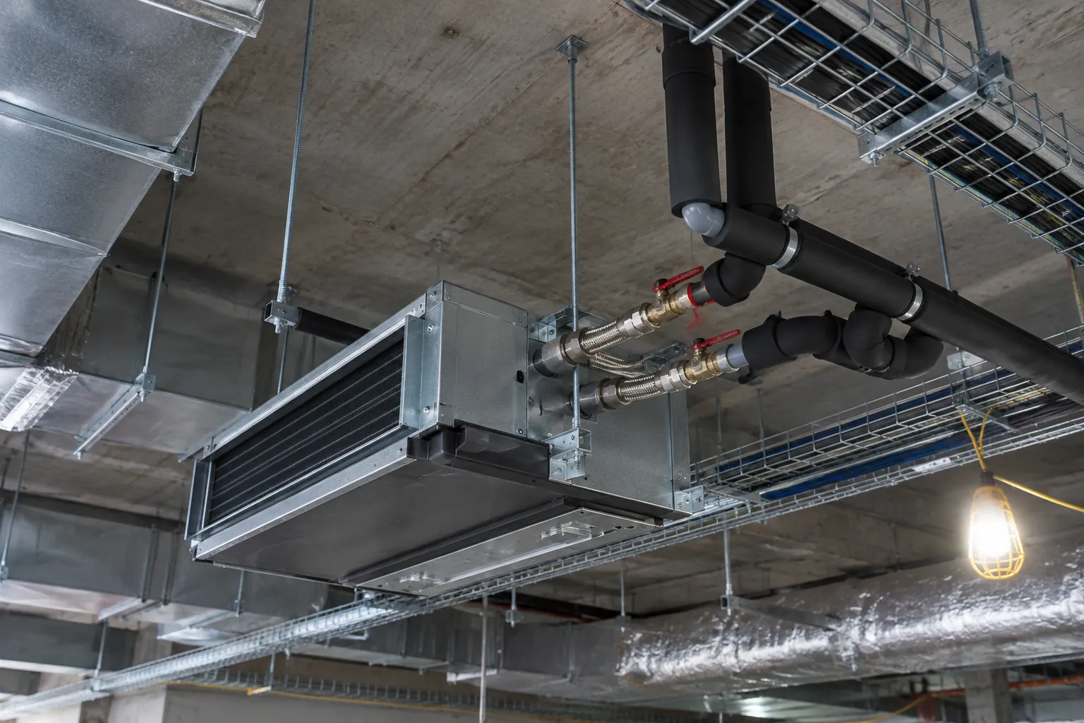 Fan-coil unit hung from a slab with insulated chilled water branch pipes, isolation valves and flexible connectors in a ceiling void